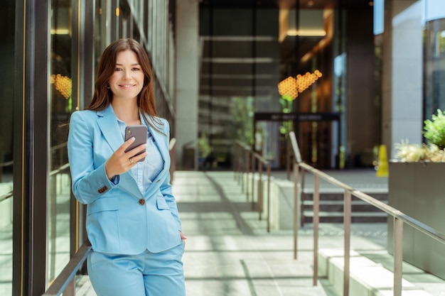 glimlachende vrouw in een stijlvol blauw zakelijk pak met een mobiele telefoon die naar de camera kijkt in het kantoor