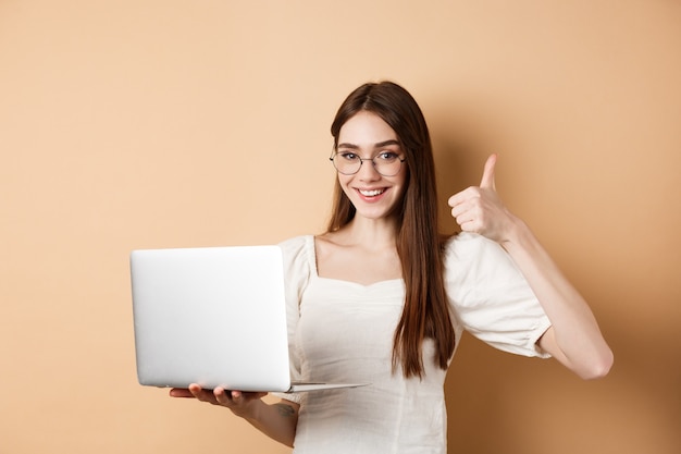 Glimlachende vrouw in glazen die laptop met behulp van en duim opdagen, die aan computer werken, die zich op beige achtergrond bevinden.