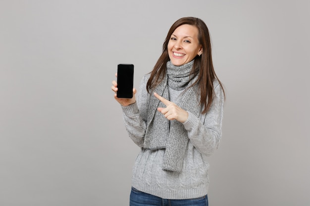 Glimlachende vrouw in grijze trui, sjaal wijzende wijsvinger op mobiele telefoon met leeg leeg scherm geïsoleerd op grijze achtergrond. Gezonde mode-levensstijl, oprechte emoties van mensen, concept van het koude seizoen.