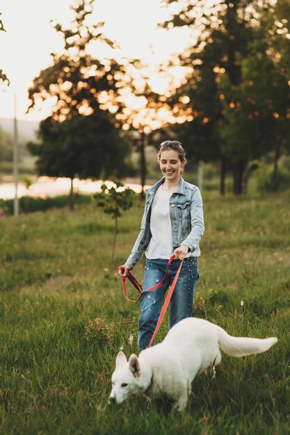 Glimlachende vrouw in jeanskleren die aan leiband witte hond op gras met vage bomen en water in avond lopen