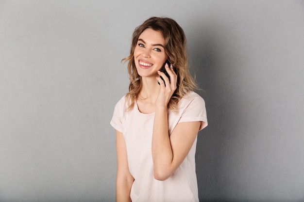 Glimlachende vrouw in t-shirt die door smartphone en over grijs spreken