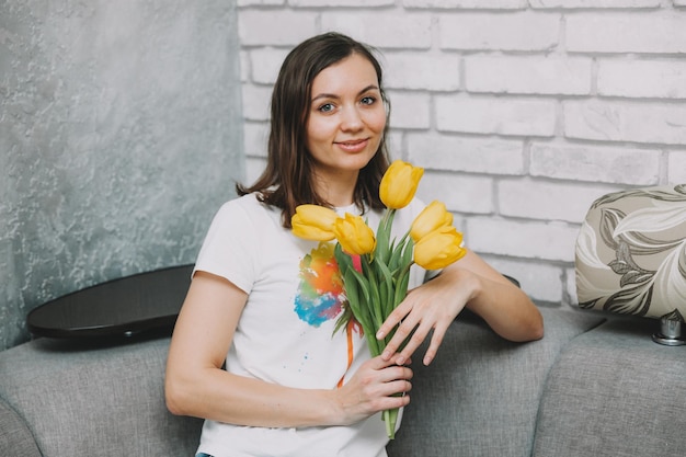 Glimlachende vrouw met gele tulpen zit thuis op de bank