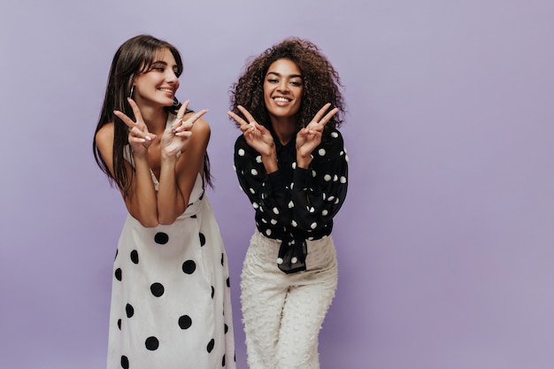 Glimlachende vrouw met golvend haar in zwarte blouse en lichte trendy broek vredestekens samen met jong meisje in moderne jurk