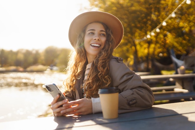 Glimlachende vrouw met hoed op het terras met telefoon geniet van zonnig herfstweer Concept van ontspanning rust