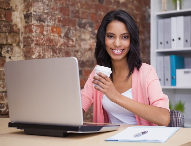 Glimlachende vrouw met koffiekop en laptop zitting bij haar bureau