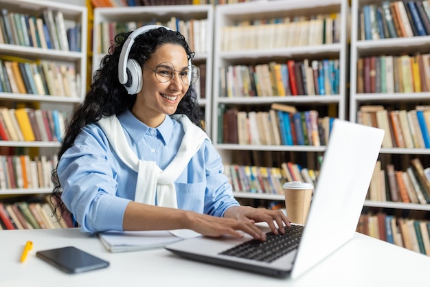 Foto glimlachende vrouw met koptelefoon die aan een laptop studeert in een bibliotheek omringd door boeken en een