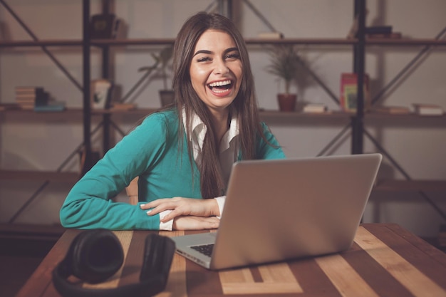 Glimlachende vrouw met koptelefoon zit aan de houten tafel met grijze laptop op tafel