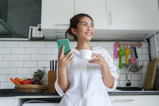 Foto glimlachende vrouw met telefoon en koffie die naar buiten kijkt in de mo