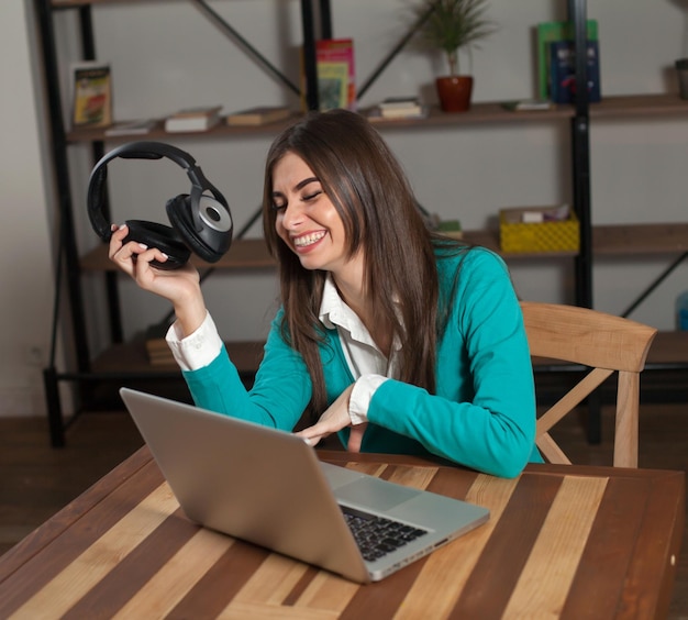 Glimlachende vrouw met zwarte koptelefoon zit aan de houten tafel met laptop