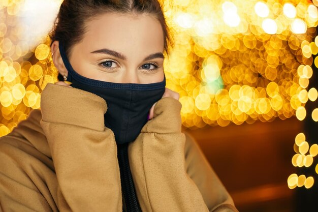 Glimlachende vrouw staat buiten 's avonds winter in vakantiesfeer en draagt een blauw gezichtsmasker