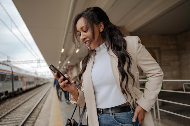 Glimlachende vrouw van gemengd ras die mobiele telefooncommunicatie gebruikt die online staat op het treinstation