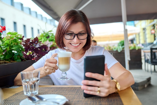 Glimlachende vrouw van middelbare leeftijd die op een terras zit met een kopje koffie en naar een smartphone kijkt