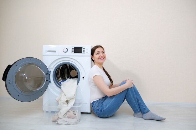Glimlachende vrouw zittend op de vloer in de buurt van moderne wasmachine met open deur en schone was in de buurt van beige muur thuis
