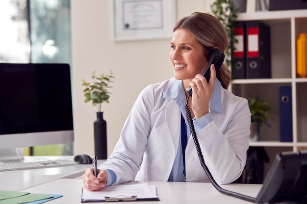 Glimlachende vrouwelijke arts of huisarts die een witte jas draagt die aan het bureau in kantoor zit en telefoneert
