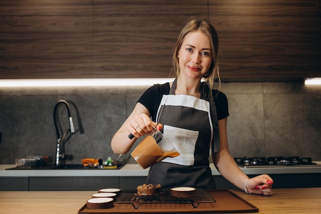 Glimlachende vrouwelijke banketbakker die vloeibare chocoladegommage op koekjescake giet Vrouwenchef-kok in uniform bij het assembleren van het proces van het moussedessert