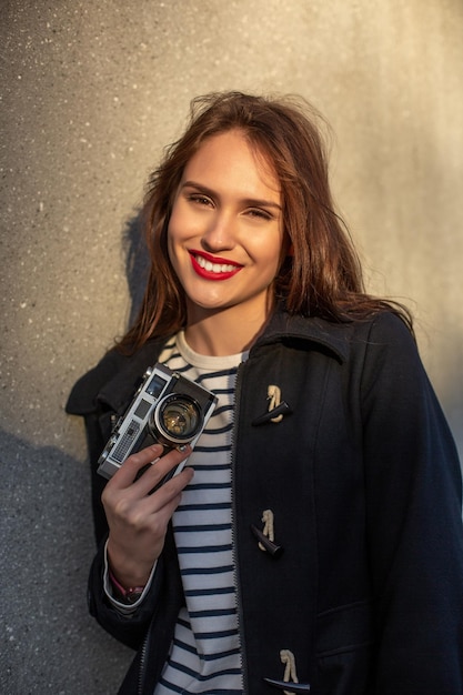 Glimlachende vrouwelijke fotograaf in jas die voor de muur staat, klaar om een nieuwe foto te maken. schattige jonge brunette vrouw in trendy outfit poseren op betonnen muur achtergrond met camera