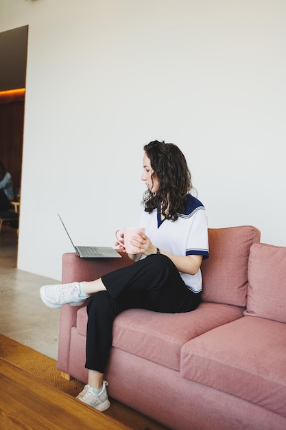 Glimlachende vrouwelijke student die op de bank zit in een helder café met koffie en naar een laptop kijkt Remote werk met een laptop in een gezellig werkcafé