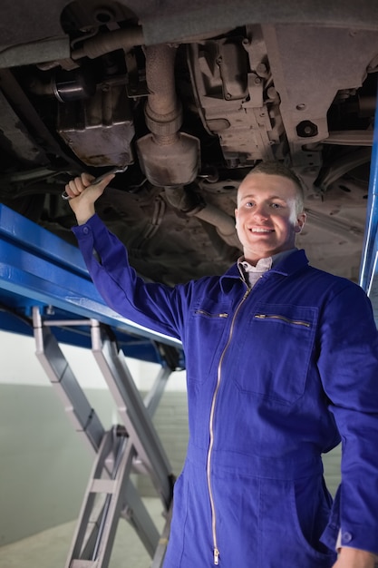 Glimlachende werktuigkundige die met een moersleutel de hieronder van een auto in een garage herstellen