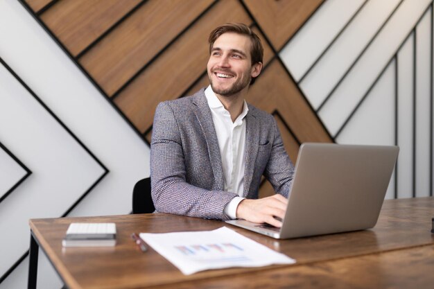 Glimlachende zakenman die aan laptop werkt en uit het raam kijkt