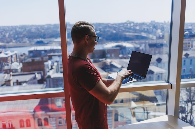 Glimlachende zakenman staat bij het raam terwijl hij aan een laptop werkt Multitasking-concept Een drukke freelancer met een bril houdt een laptop in zijn handen terwijl hij bij het raam staat Smart casual