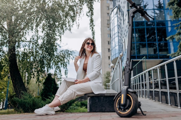 Glimlachende zakenvrouw in wit pak en zonnebril bezig met digitale tablet zittend in de buurt van modern kantoorgebouw met elektrische scooter op de voorgrond