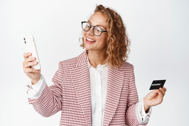 Glimlachende zakenvrouw met een bril, creditcard vast, kijkend naar haar mobiele telefoon, online winkelen op smartphone-app op wit.