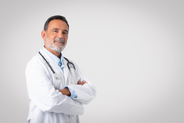 Foto glimlachende zelfverzekerde blanke senior arts-therapeut in witte jas met gekruiste armen geniet van werk