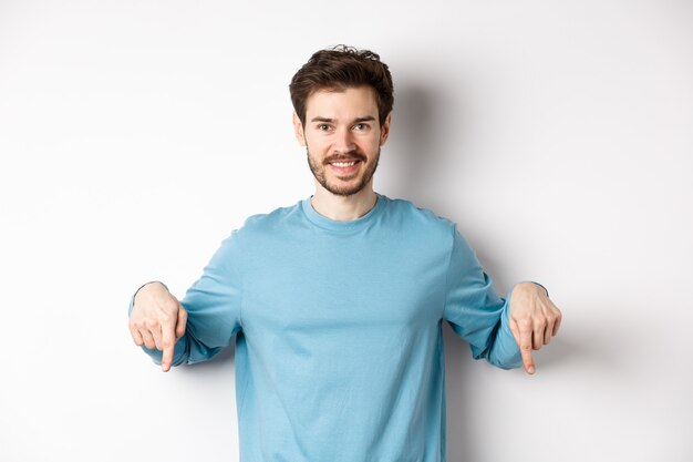 Glimlachende zelfverzekerde man die met zijn vingers naar beneden wijst, promobanner of logo op een witte achtergrond toont, staande in een casual blauw sweatshirt