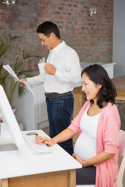 Glimlachende zwangere vrouw die laptop thuis met behulp van terwijl haar echtgenoot krant leest