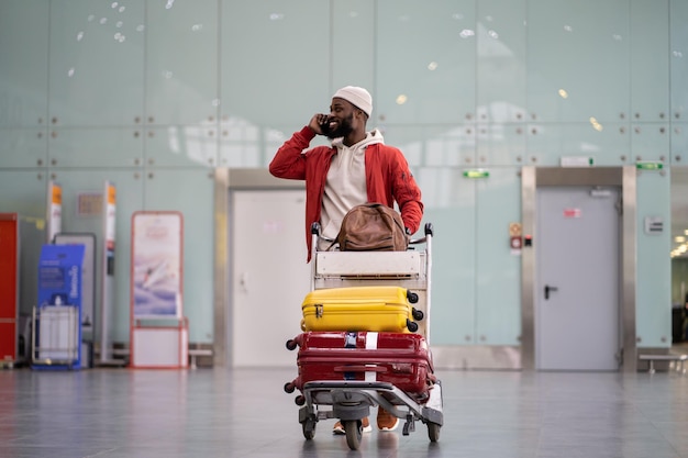 Glimlachende zwarte man die bagagewagen duwt die loopt na aankomst op de luchthaven terwijl hij op mobiele telefoon praat