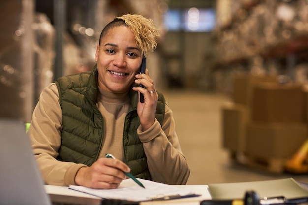 Glimlachende zwarte vrouw die telefonisch spreekt in het magazijn en naar de camera kijkt