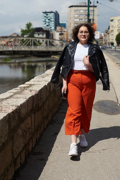 Glimlachende zwarte vrouw in de kleding van de stylist en een bril die geniet van een zonnige dag Collegemeisje dat over de stad loopt