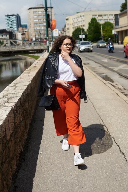 Glimlachende zwarte vrouw in de kleding van de stylist en een bril die geniet van een zonnige dag Collegemeisje dat over de stad loopt