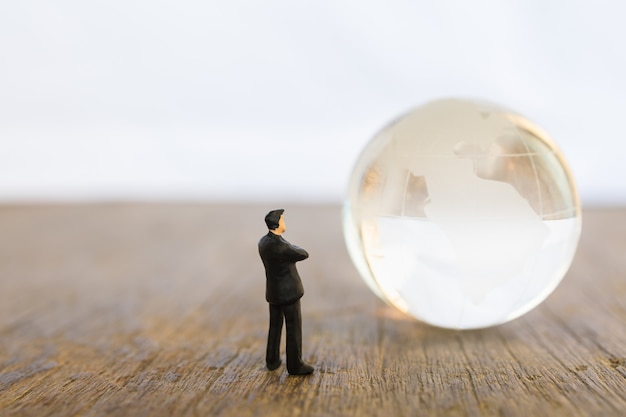 Global and Business Concept. Businessman miniature people figure standing and looking to mini world glass ball on wooden table with copy space.