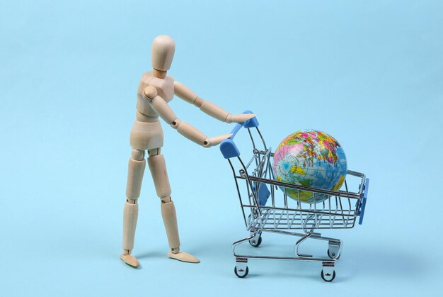 Photo global shopping concept. wooden puppet with supermarket trolley and globe on blue background