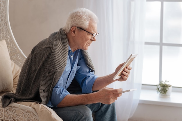 Gloomy depressed man looking at the old photograph and remembering his past while sitting near the window