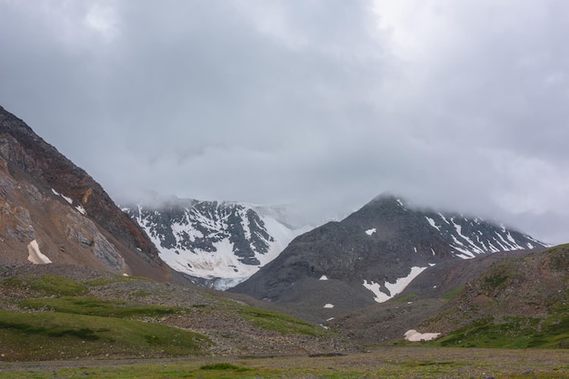 Gloomy landscape with snow mountain range in gray low clouds in overcast Atmospheric mountain scenery with snow mountains among low clouds Dramatic view to mountains under gray sky in rainy weather