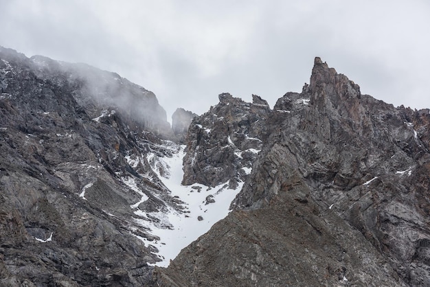 写真 灰色の低い雲に雪が降る高い鋭いロッキーのある暗い山の風景 鋭い岩と灰色の曇り空にある尖った頂上のクローズアップ 先のとがった岩のある山脈のある荒涼とした曇りの風景