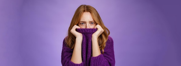 Gloomy upset redhead girlfriend pulling collar of purple sweater on nose frowning and squinting offe