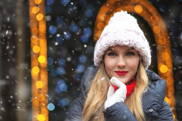 Glorieuze blonde vrouw draagt een gebreide muts die tijdens de sneeuwval op de wintermarkt loopt. Lege ruimte voor tekst