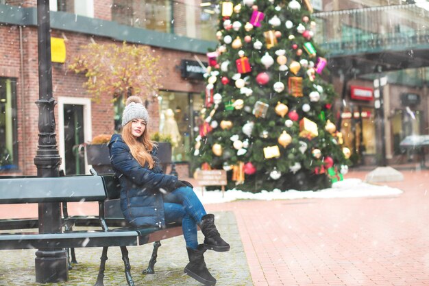 Glorious redhead woman walking near decorated new year tree during snowfall. Space for text