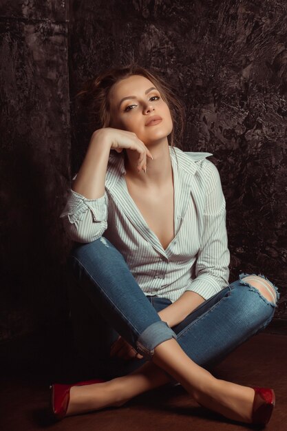 Glorious young woman in shirt and jeans sitting on the floor at studio