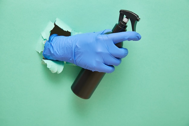 A gloved hand holds a disinfectant in a black spray bottle. Green paper background with torn hole