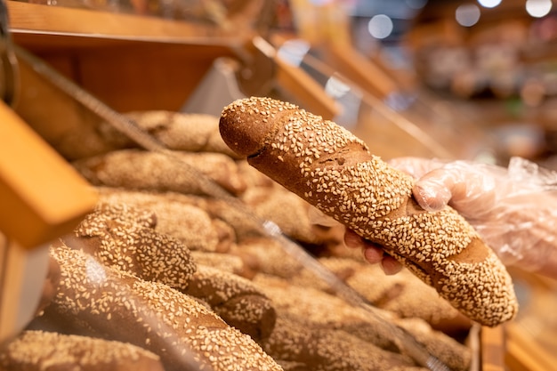 Mano guantata del compratore femminile maturo che tiene pane fresco cosparso di semi di sesamo mentre è in piedi dal display nel supermercato