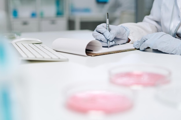 Gloved hands of biochemist writing down results of scientific experiment