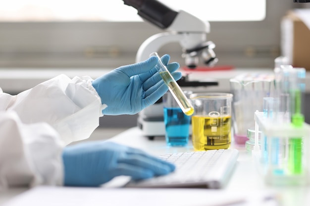 Gloved scientist holds test tube with transparent liquid and types on keyboard chemical