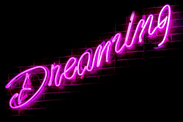 Фото Светящаяся фиолетовая неоновая надпись dreaming на фоне темной кирпичной стены. винтажное изображение темных тонов.