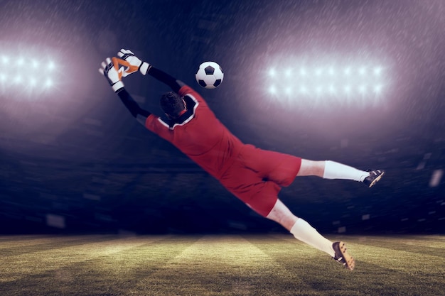 スポット ライトの下でサッカーのピッチに対して赤でジャンプ アップのゴールキーパー