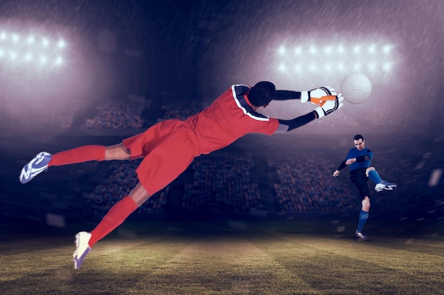 Goalkeeper in red making a save against large football stadium under blue sky