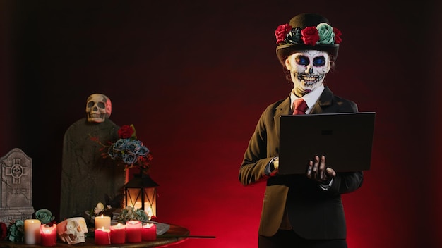Goddess of death surfing internet with laptop on dios de los muertos mexican ritual, celebrating holy santa muerte with suit and hat halloween costume. Looking at website on wireless pc.